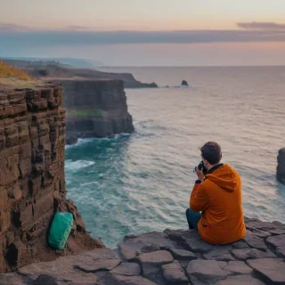 A serene oceanside cliff of basalt columns in bokeh photography style, using only the colors orange, purple, teal, and green, with the sky visible and a subtle gradient of colors to give a sense of depth, a person sitting peacefully on the edge of the cliff, taking a photo of the sunrise with a camera