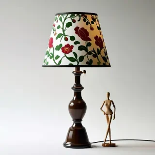 A lamp with a floral patterned lampshade. There is a tiny mannequin about half the size of the lamp leaning against the lamp in a casual position