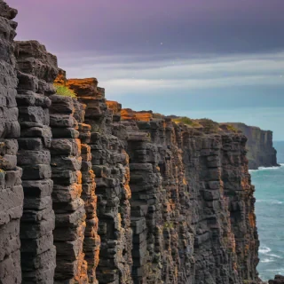 An oceanside cliff of basalt columns in bokeh photography style, using only the colors orange, purple, teal, and green, with the sky visible and a subtle gradient of colors to give a sense of depth.
